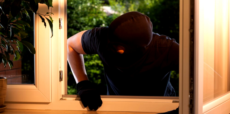 burglar wearing black gloves and a black mask breaking into a home through a window